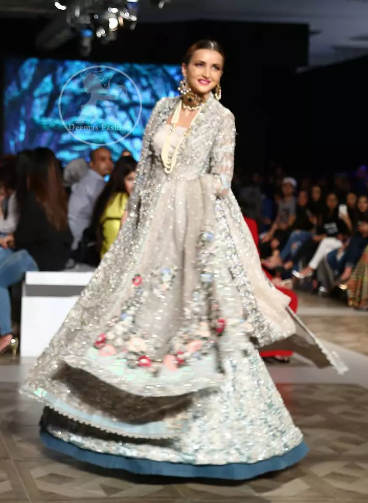 This outfit is a timeless beauty. It is heavily embellished with silver gold kora dabka, Sequins and swarovski crystals. This exquisite Pishwas is fully decorated with floral motifs patterns all over it. It is further enhanced with multiple colored Foral thread embroidery. The applique border of pishwas is ornamented with small silver pearls. It comes with embellished lehenga which has small sized sprinkled floral motifs all over. This Outfit is beautifully coordinated with matching Dupatta with heavy embroidered borders.