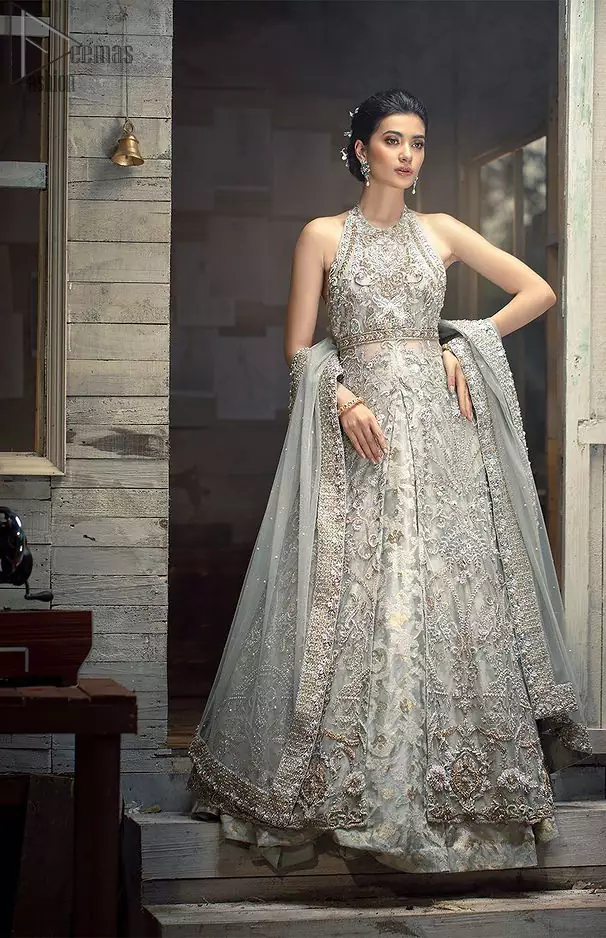 Nikah Wear - Light Grey Halter Neck Gown Lehenga. The floral and the flowery climber networks on fully embellished Halter neck and blouse has exquisitely exaggerated in the beauty of the costume making it very unique. The bottom lehenga with Fixed Waist Belt With Side Zip Closure is composed of Katan Banarsi Jamawar fully embellished with flowery climbers with Light Gray colour tone; being exquisitely beautiful in perfecting the complete outfit having a unique fabric and double flare lehenga that is in stellar combo with the blouse with finished edges and Dupatta sideways carried beautifully by the bride with organza fabric and Heavy beautiful golden and Grayish lace on all four bordered finished sides and dabka work on the net inside of the gorgeously made dupatta, whole this costume has its own unifying feature such as 100 % imported high quality overall highly embellished fabric making it the best choice to have by you on your big day.