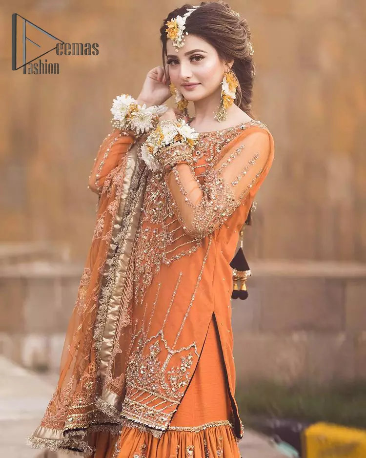 Time to treat yourself with an orange article of Deemas Fashion. This orange heartiest mealtime article will twirl your heart.  Prettify your Mehndi look with this orange short shirt that is embellished with tilla, kora and dabka work. Its rounded neckline with full sleeves gives you a genuine feeling of a bride. It is synchronized with flayer orange sharara that has an embellished border and spray of tiny floral motifs that gives a super aesthetic look to this dress. Finish this look with a net dupatta that has four sides embellished border and Kiran work.